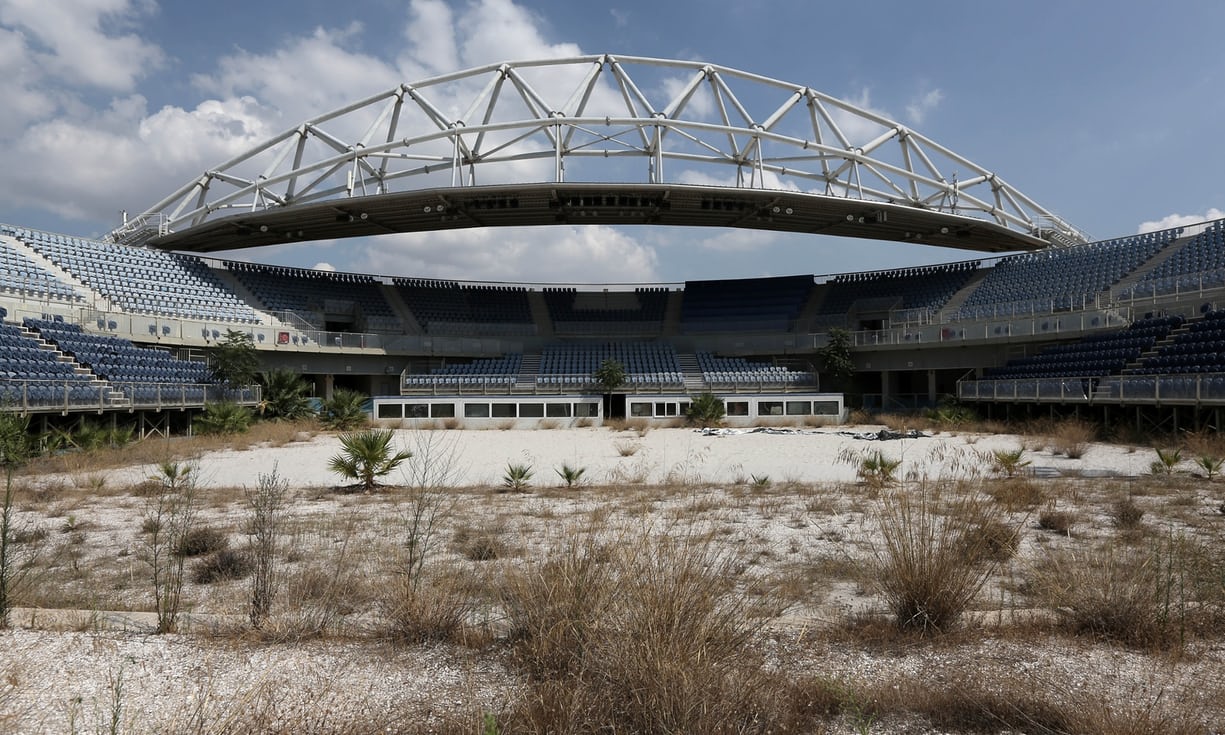 An abandoned beach volleyball stadium in Greece. Weeds grow through the sand on the court, the permanent steel and concrete stands sit vacant.