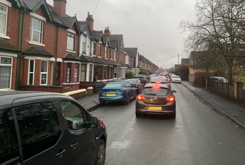 A residential street is shown with parked cars along both sides and a line of stationary traffic in the middle. The road is completely blocked.